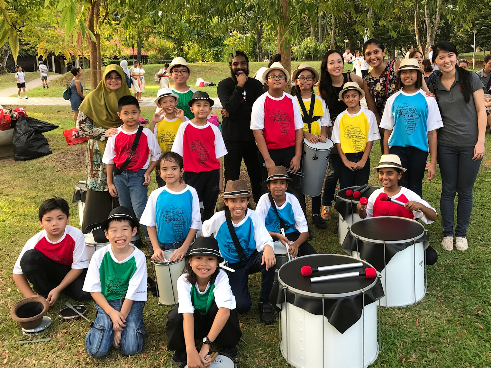 A group of children in colorful percussion band uniforms and hats pose outdoors with drums and adult chaperones.